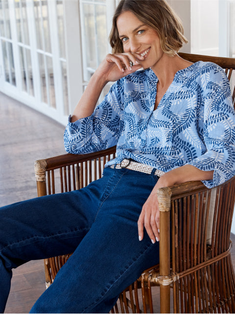 Woman sitting on a wooden chair wearing a blue patterned shirt and jeans.