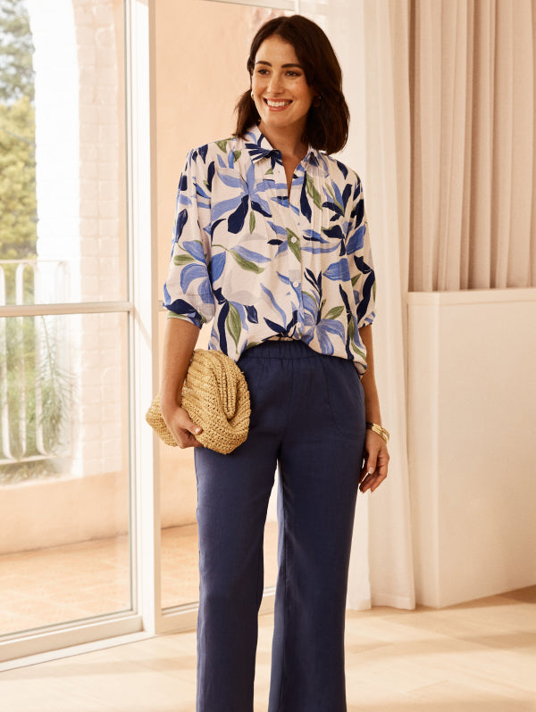Woman wearing a floral blouse and navy pants in a bright room.